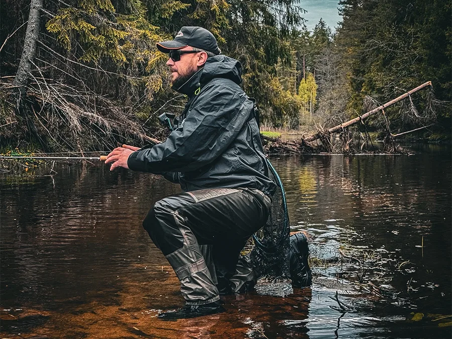 Una persona con vadeadores pescando en un río de bosque entre árboles
